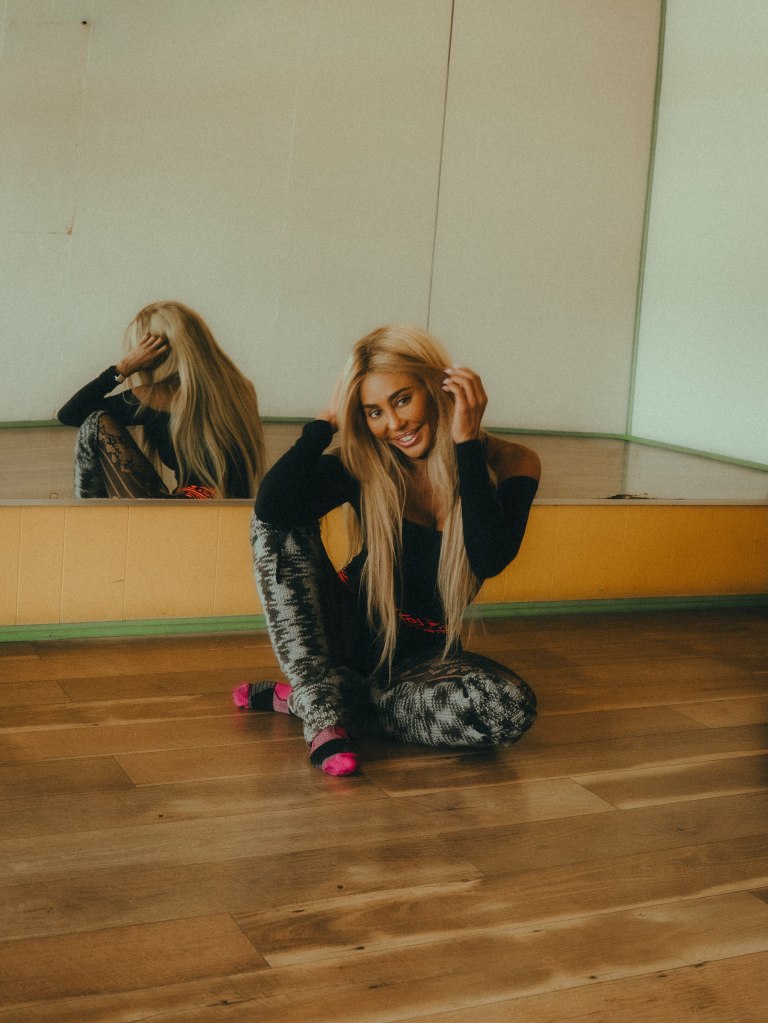 Singer Shyra Sanchez sitting on a wooden floor in a dance studio with her reflection visible in the mirror behind her.