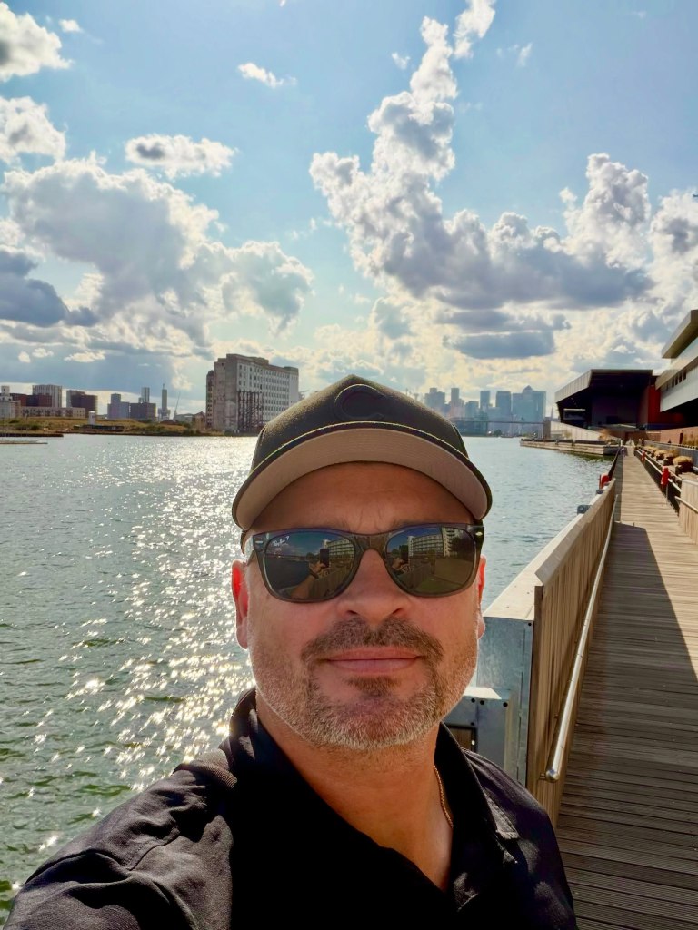 Tom Welling takes a selfie wearing a baseball cap and sunglasses on a boardwalk next to a body of water with a city skyline in the distance.