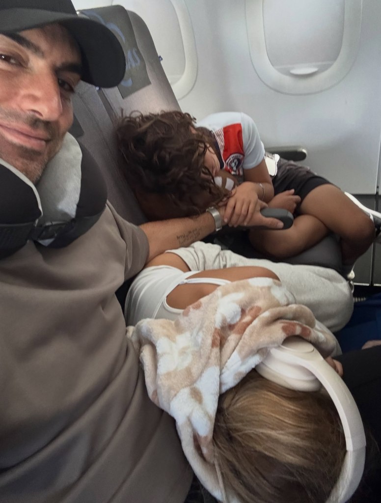 Justin Baldoni smiling for a selfie on an airplane while two children sleep comfortably next to him.