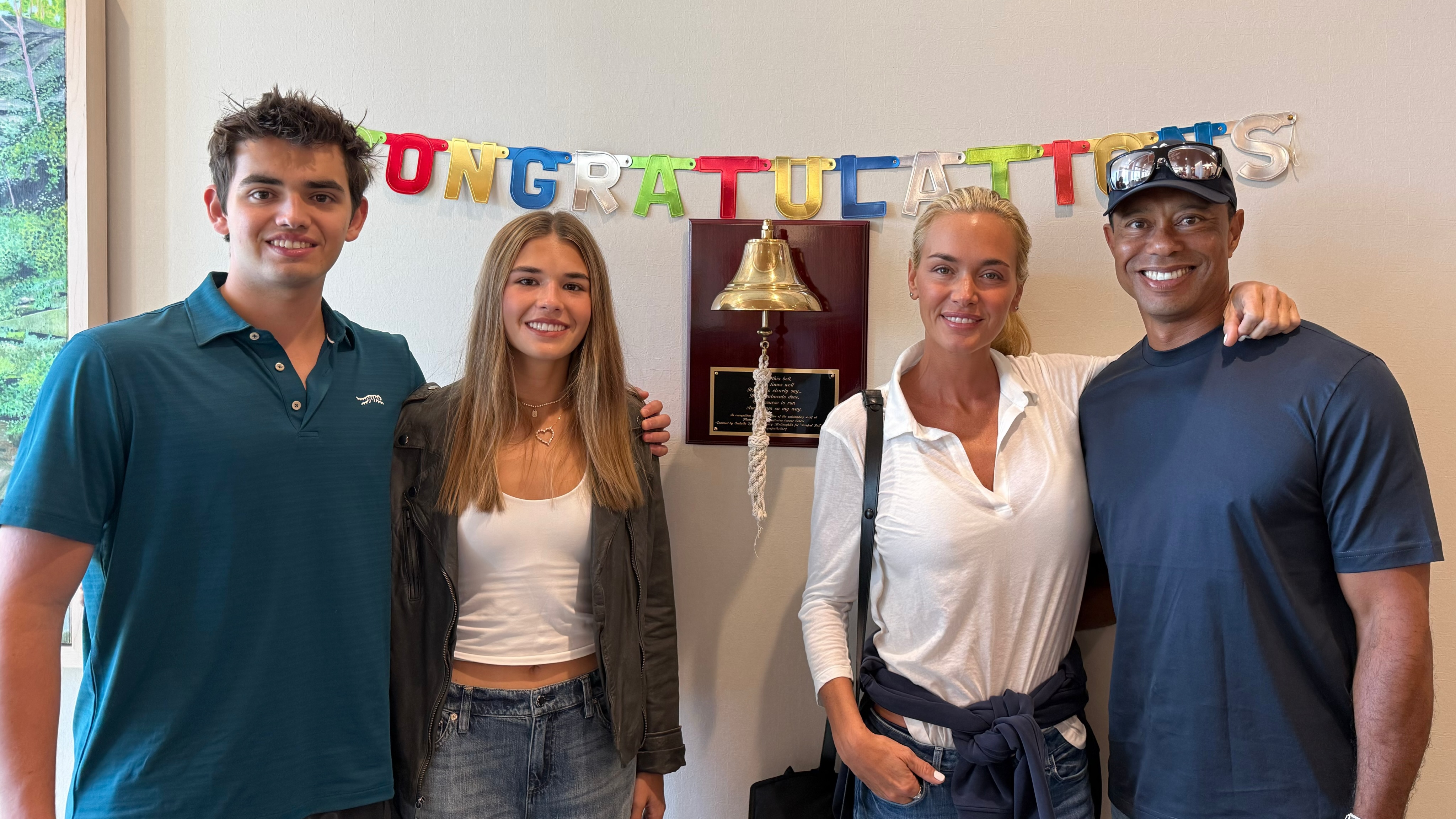 Vanessa Trump, Kai Trump, and Tiger Woods pose together in front of a congratulatory banner and a mounted bell.