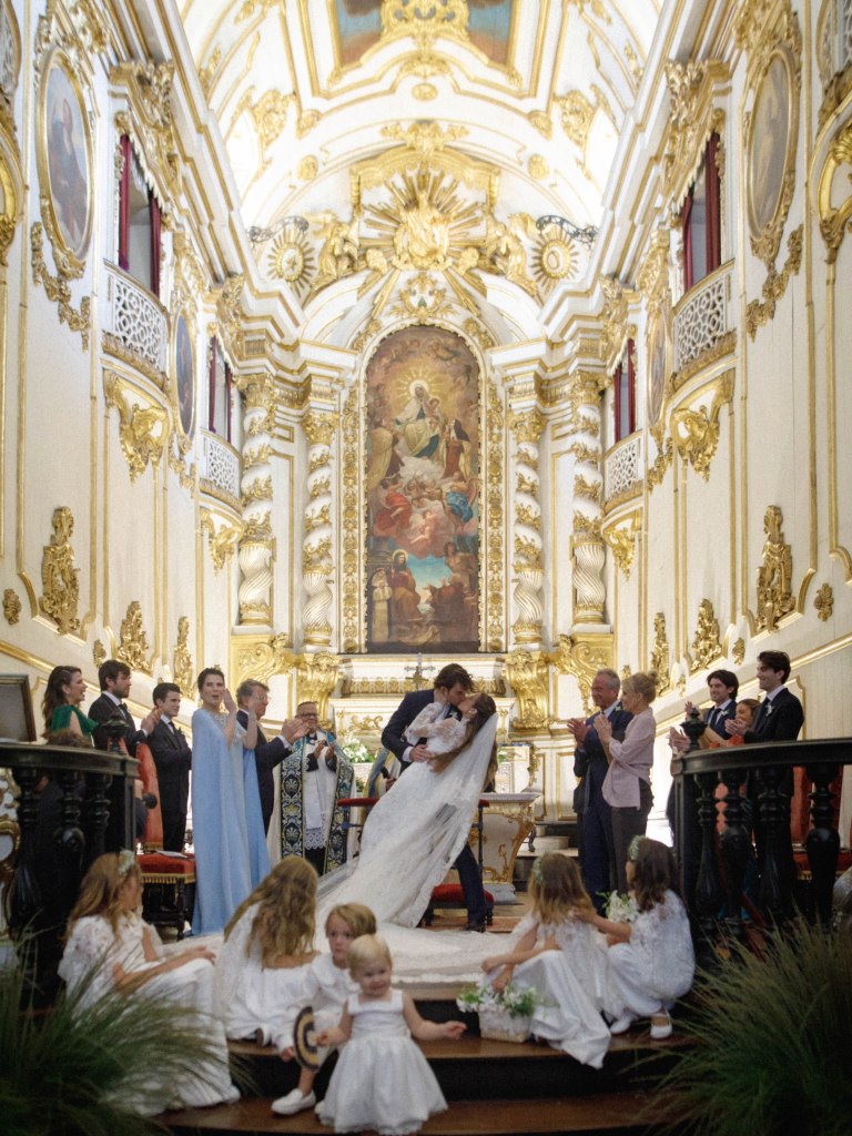 Conor Kennedy and his bride kiss at the altar of a church during their wedding ceremony, surrounded by guests and flower girls.