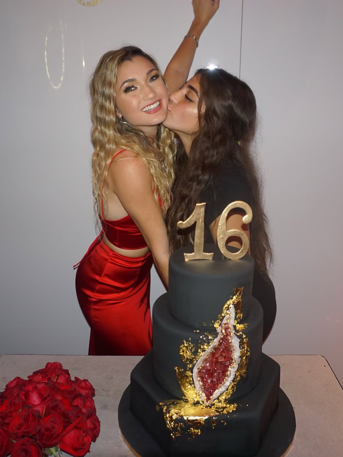 Milania Giudice kisses sister Gabriella on the cheek next to a black and gold birthday cake with the number 16 on top.