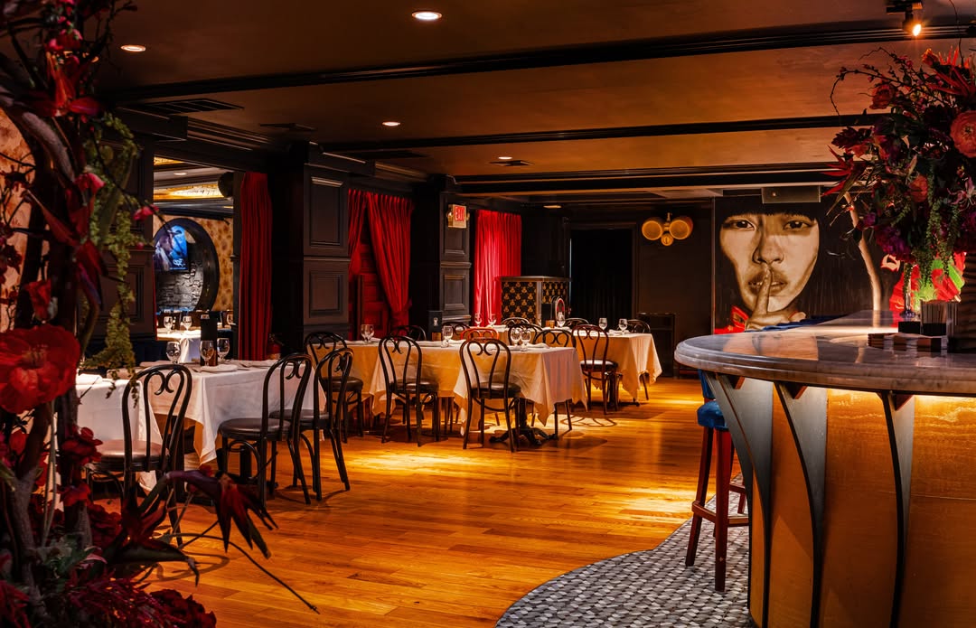 Interior view of a dimly lit, elegant restaurant featuring wooden floors, white-clothed dining tables with dark chairs, and a bar counter in the foreground.