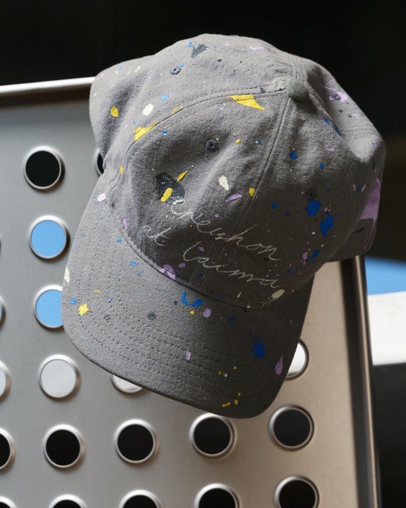 A gray Erewhon hat with paint splatters hanging on a metal surface.