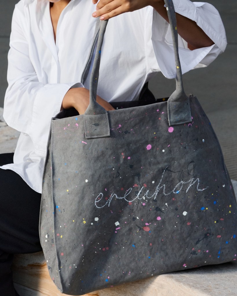 A person wearing a white shirt holds a dark grey Erewhon tote bag decorated with colorful paint splatters.