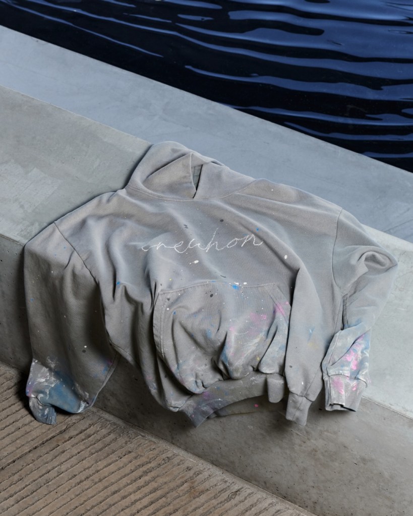 A grey hoodie with paint splatters and the Erewhon logo, resting on a concrete ledge near water.