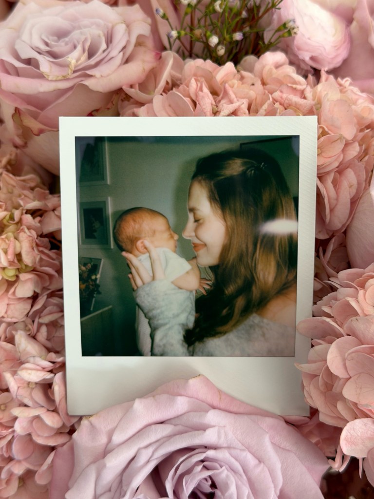 A Polaroid photo of a woman holding and gazing at a newborn baby, resting on a bed of pink flowers.
