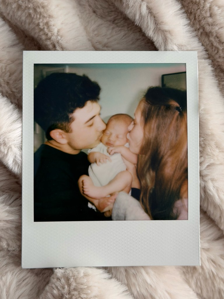 A Polaroid photo of a man and a woman kissing a sleeping newborn baby they are holding.