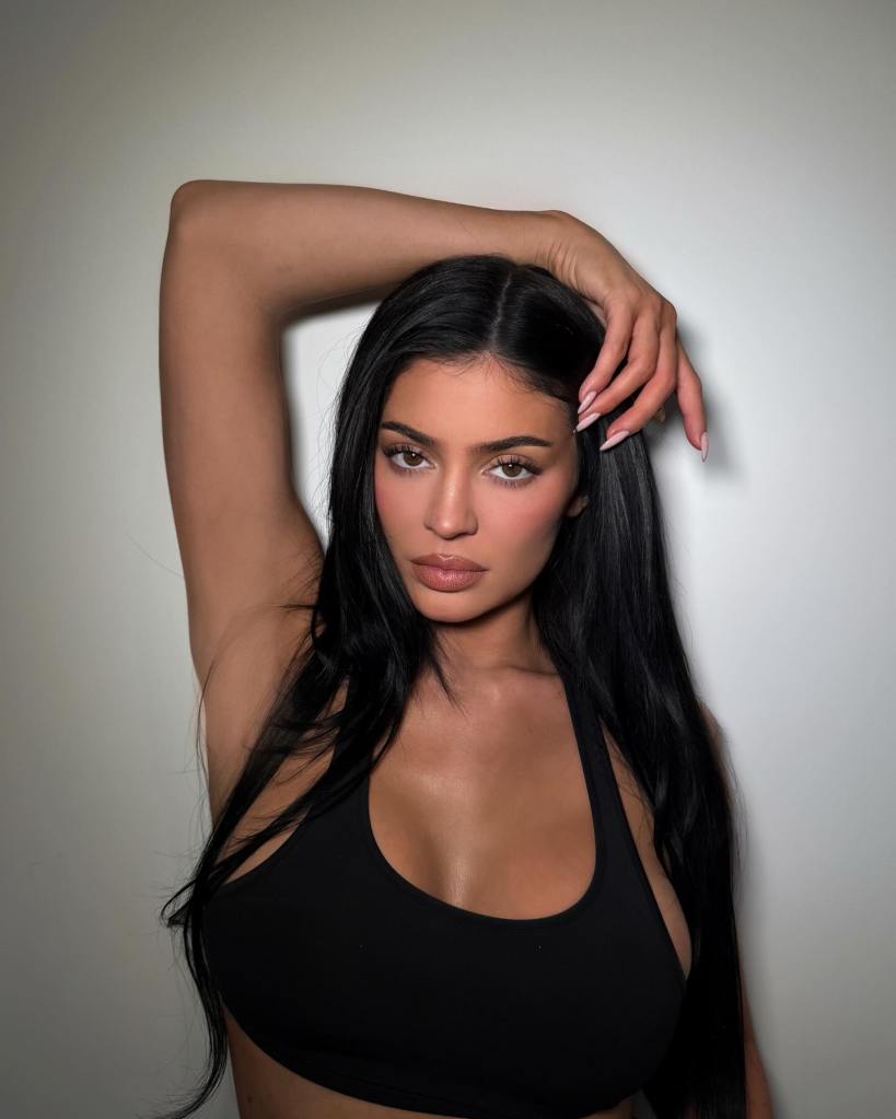 Kylie Jenner poses with her right arm raised, hand touching her long black hair, wearing a black top against a plain wall.