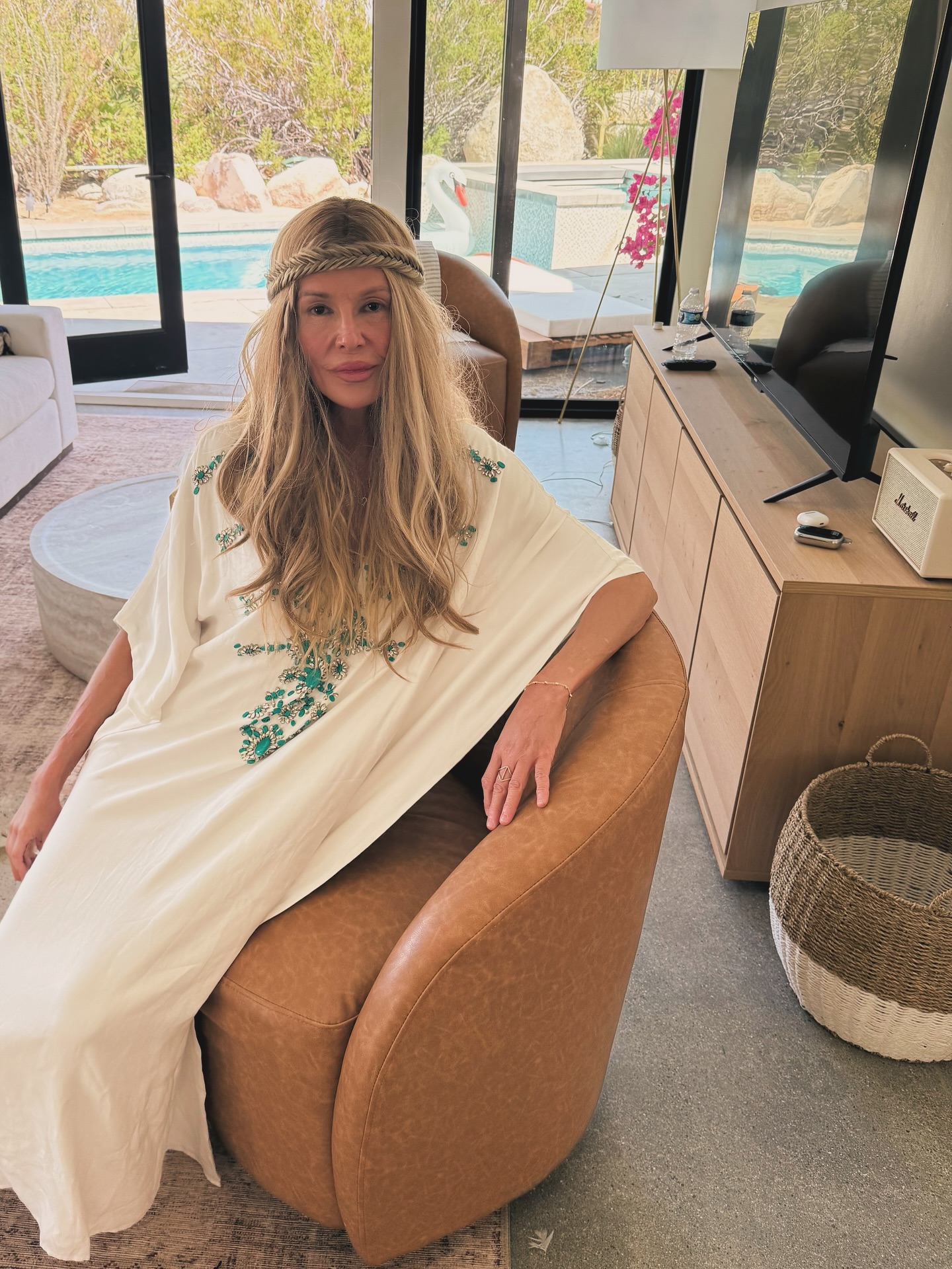 Brandi Glanville sits in a leather armchair wearing a white kaftan and a braided headband in a living room with a pool view.