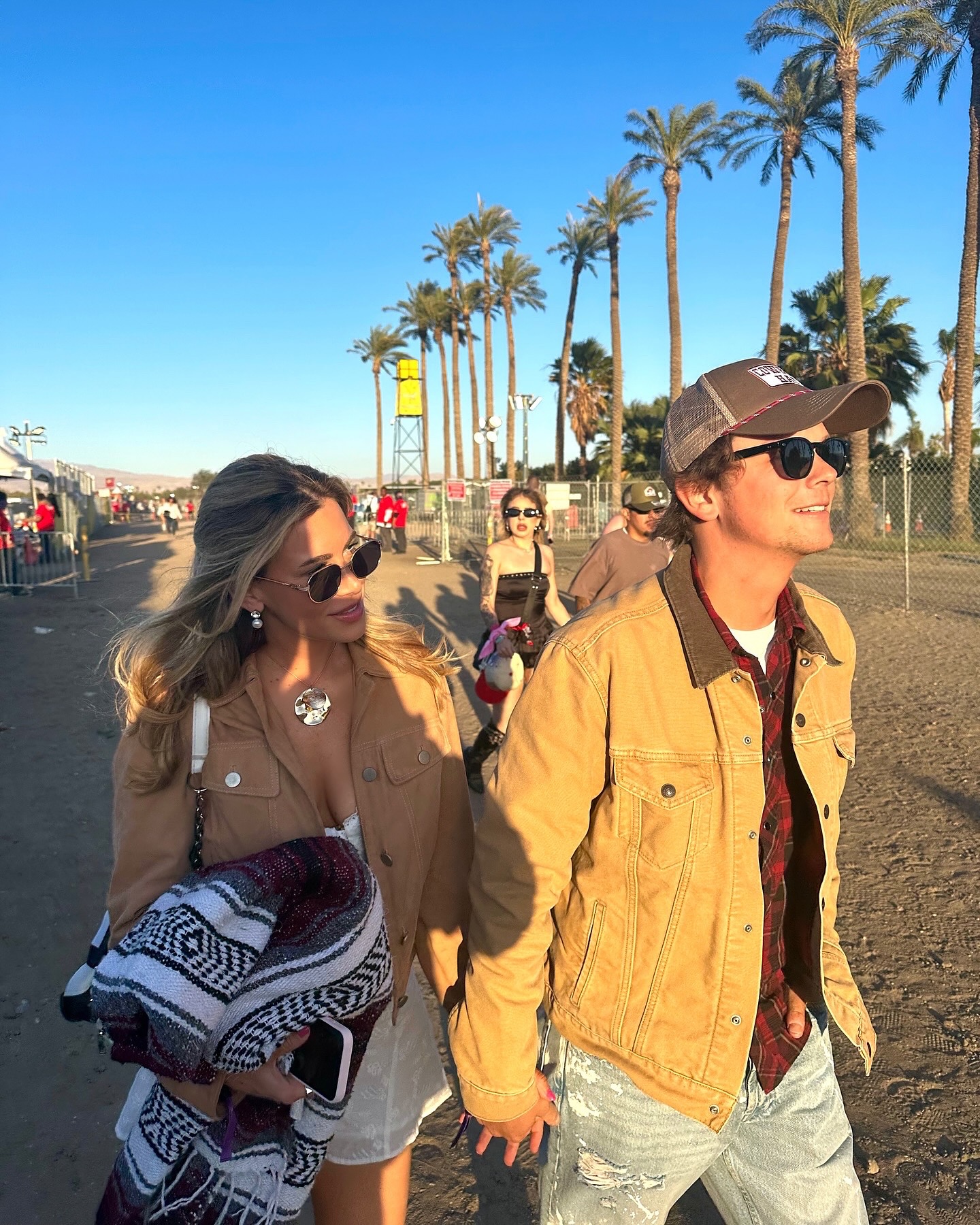 Matthew Lowe and Emily Markus walk together outdoors, holding hands near a line of palm trees.