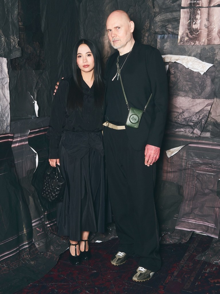 Billy Corgan and Chloe Mendel pose together in front of a textured backdrop, both dressed in dark clothing.