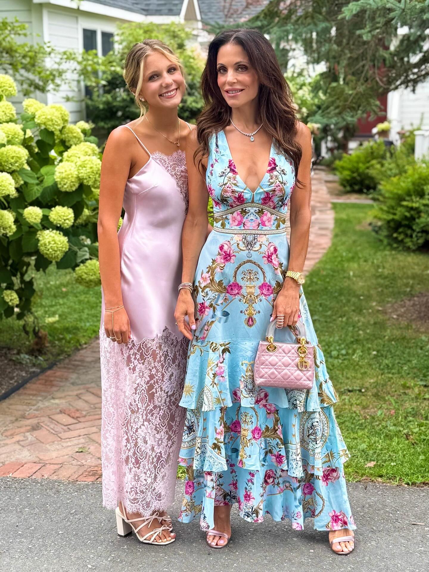 Bethenny Frankel and her daughter Bryn Hoppy pose together outdoors, with Frankel in a light blue floral dress and Hoppy in a pink satin dress with lace trim.