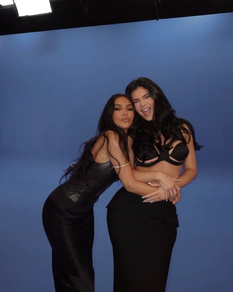 Kim Kardashian and Kylie Jenner pose together in black outfits against a blue backdrop.