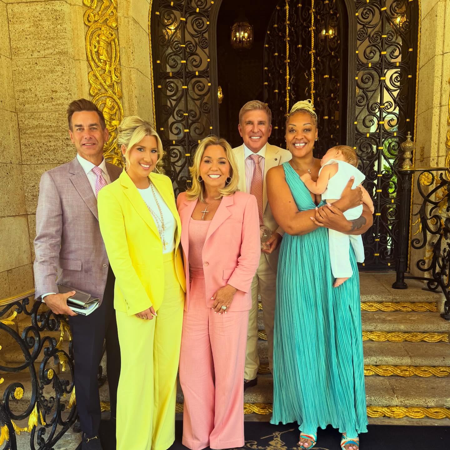 The Chrisley family posing with Savannah Chrisley's fiancé, Jason, and another woman holding a baby in front of a grand doorway.