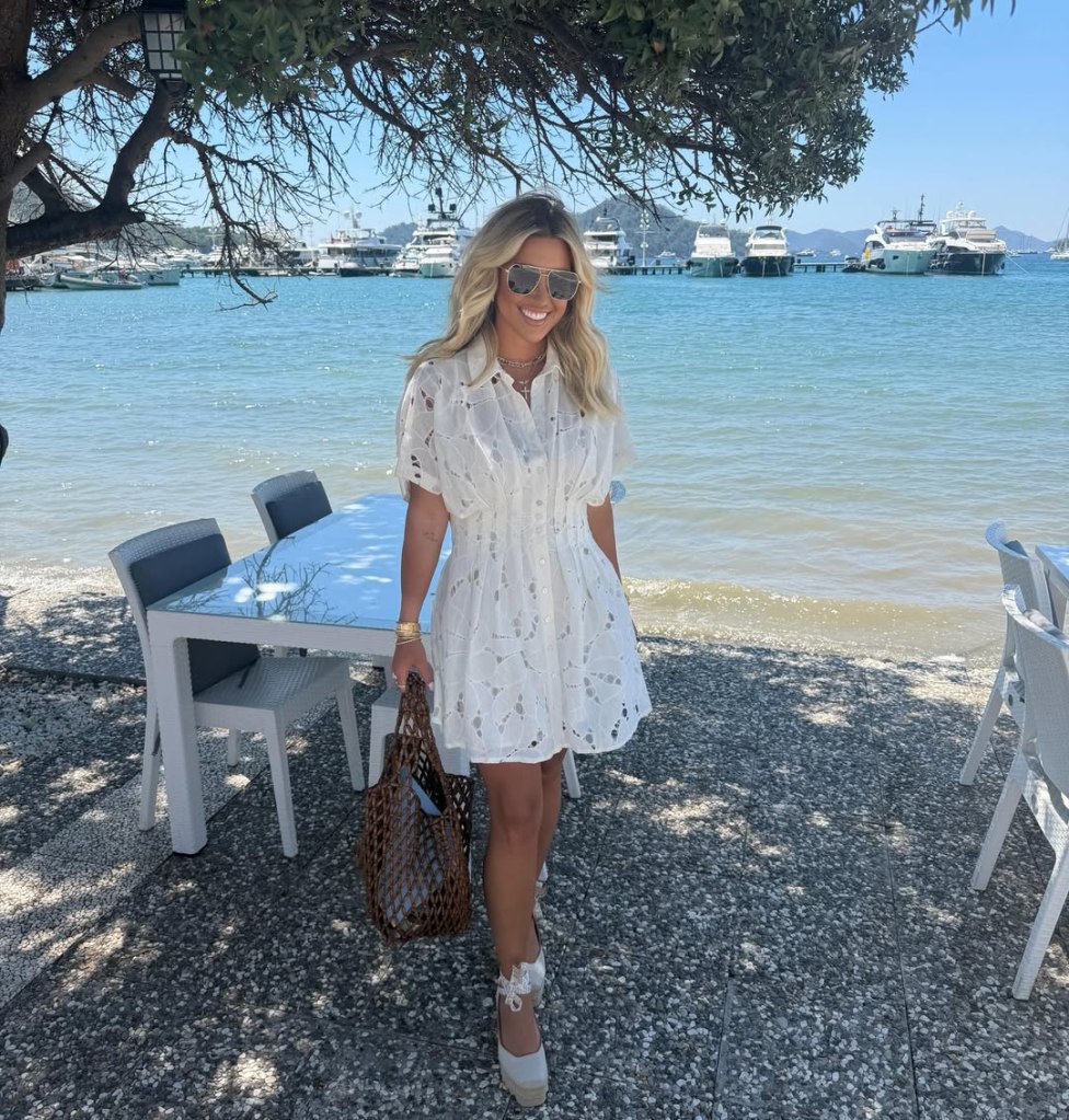 Savannah Chrisley stands by a waterfront restaurant holding a brown woven bag, wearing a white lace dress and sunglasses.