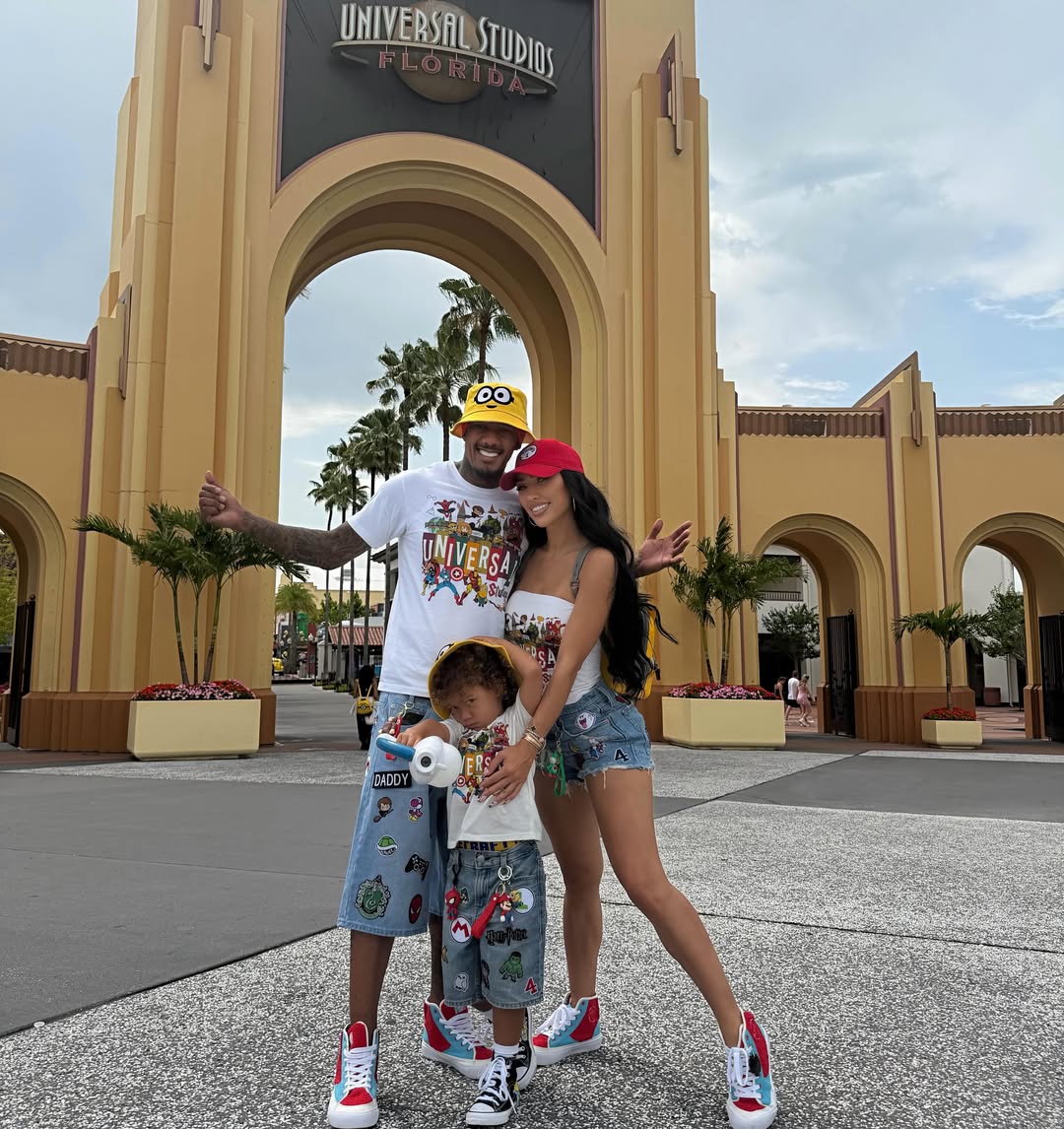 Nick Cannon and Bre Tiesi stand together with their child in front of the Universal Studios Florida entrance archway.