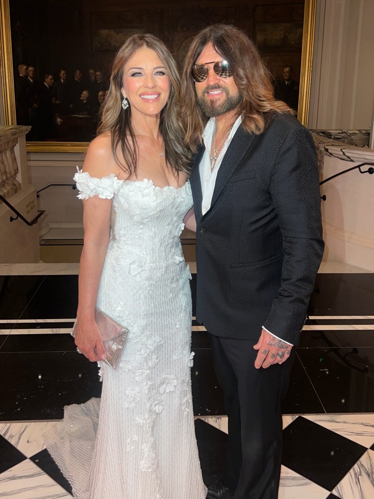 Elizabeth Hurley in a white off-the-shoulder gown standing next to Billy Ray Cyrus in a black suit.