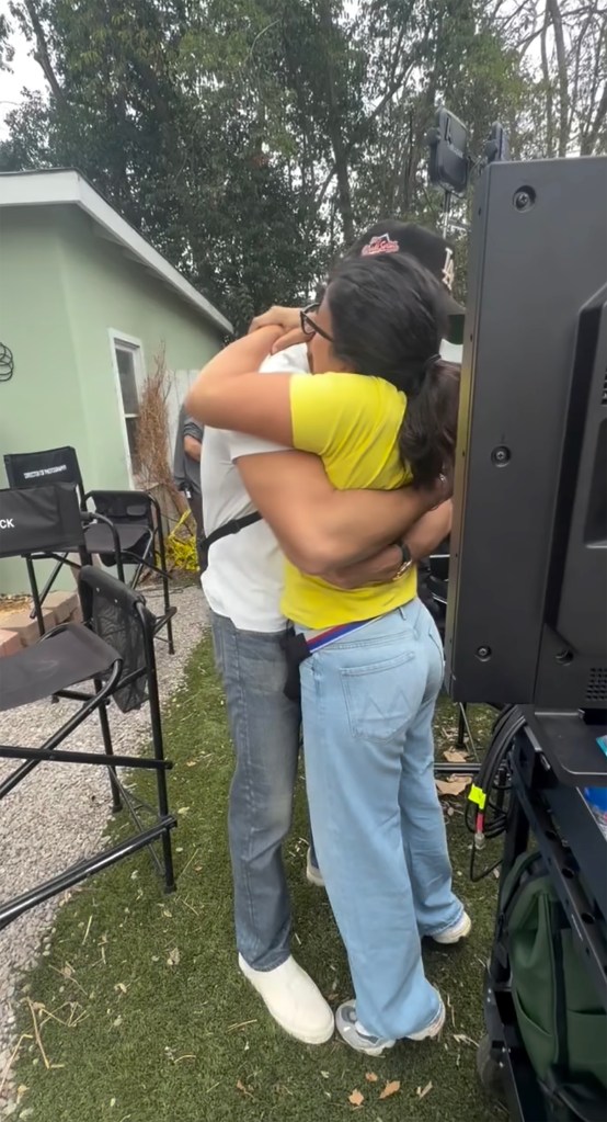 Two people are hugging outdoors on a film set, with director's chairs and a monitor visible in the background.