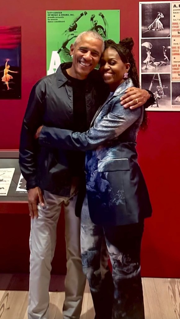 Barack and Michelle Obama embrace and smile in front of a red wall with posters.