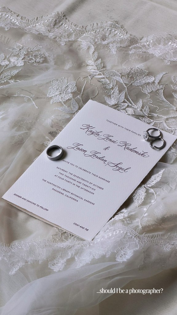 Wedding invitation for Kayla Radomski and Jason Segel, resting on a lace fabric with wedding rings.