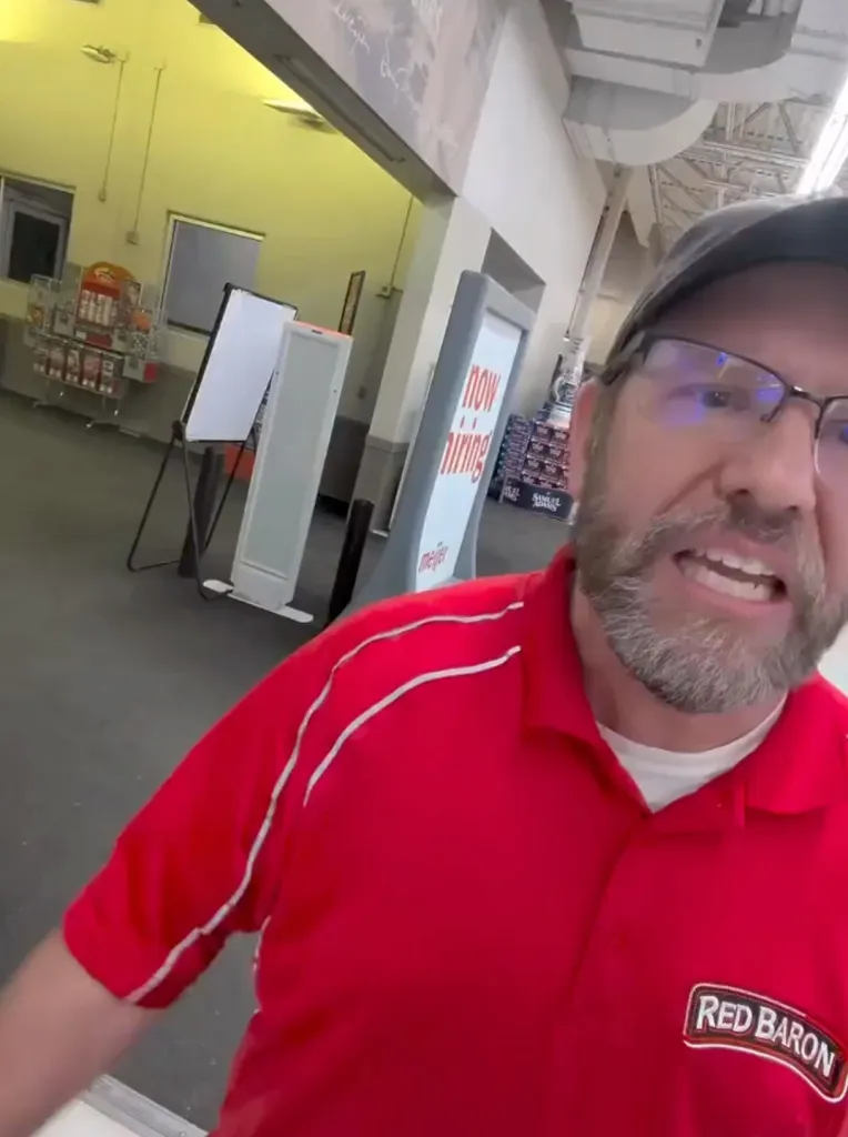 A man wearing glasses and a red polo shirt with the Red Baron logo is speaking to someone while standing inside a retail store.
