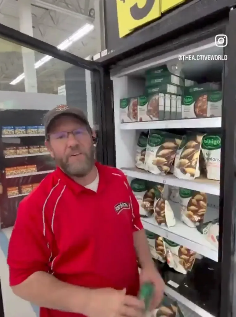 A man in a red polo shirt and baseball cap stands in front of a grocery store freezer.