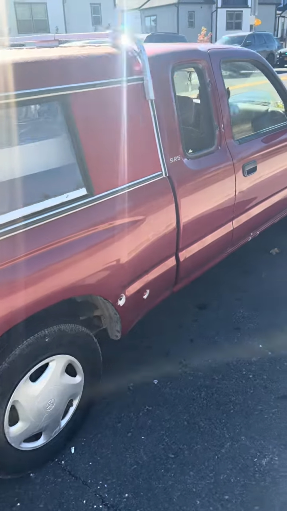A red pickup truck with bullet holes on the side, parked on a street.