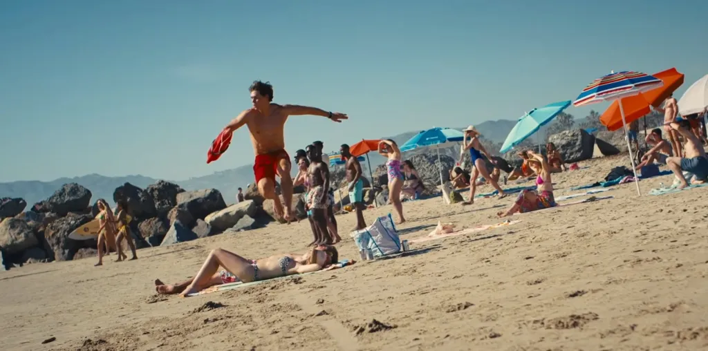 A shirtless lifeguard leaps over a sunbather on a busy beach with colorful umbrellas.