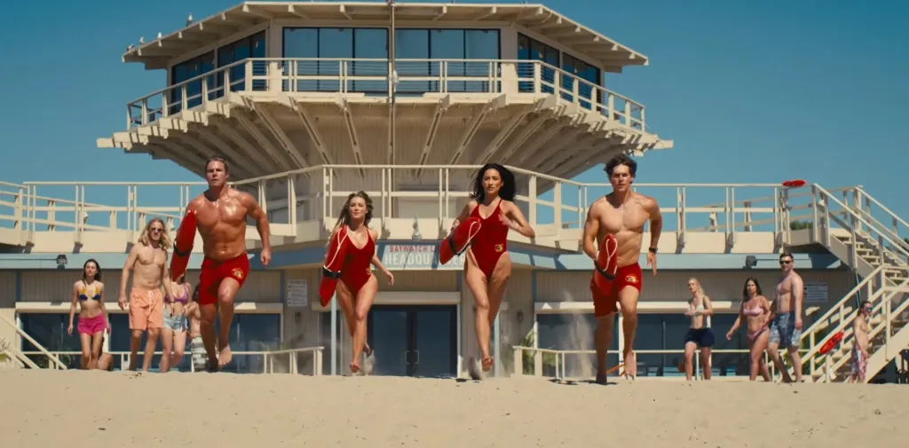Baywatch lifeguards running on the beach in slow motion, in front of a lifeguard tower.