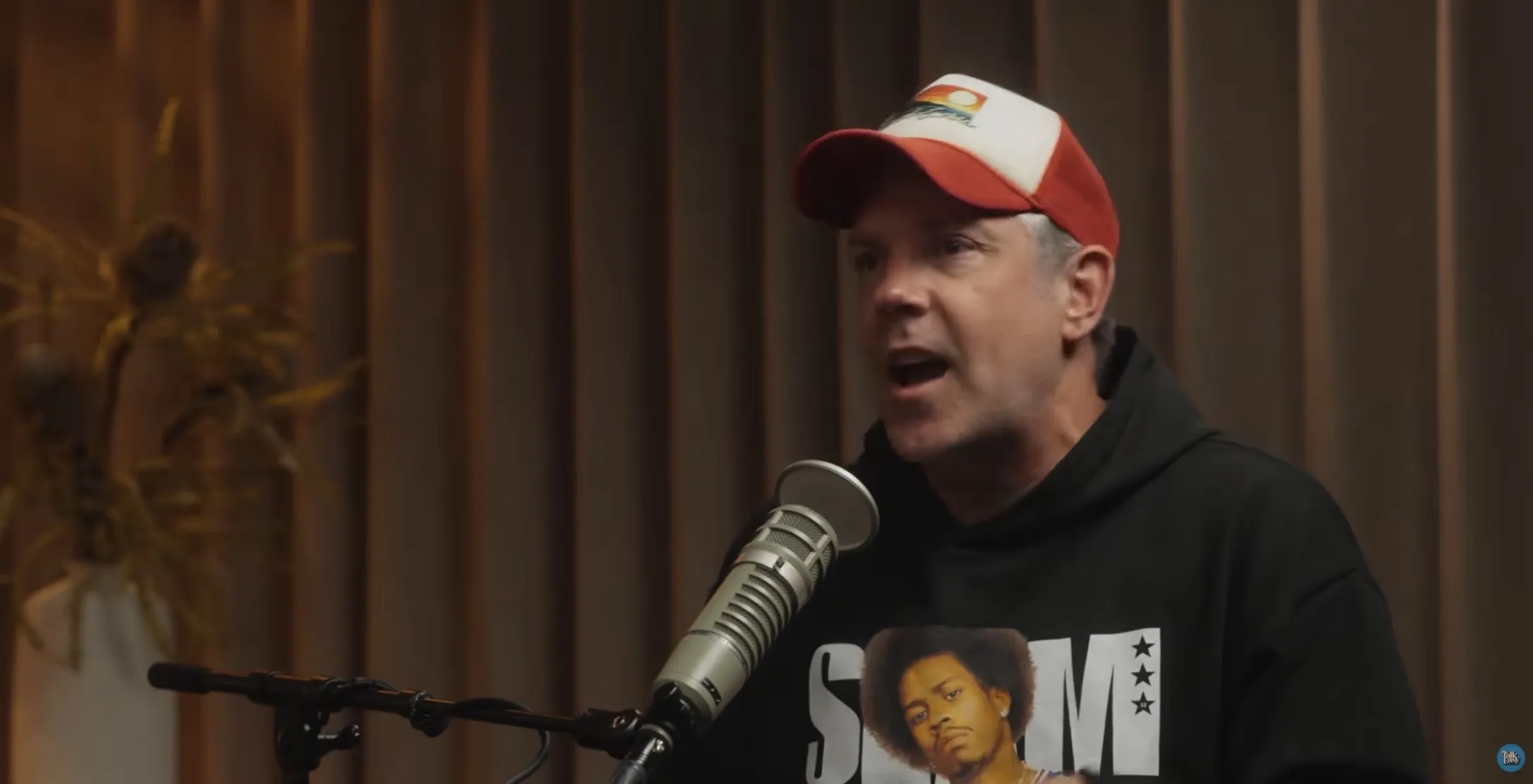 Jason Sudeikis speaking into a microphone while wearing a baseball cap and a black hoodie.