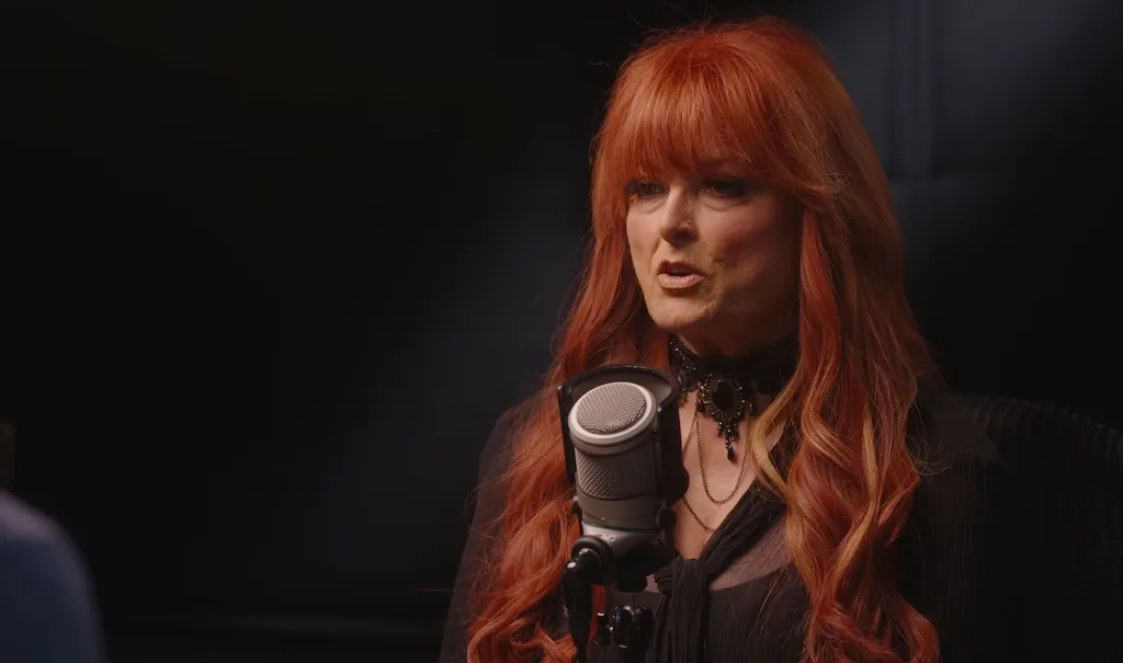 Wynonna Judd with red hair and a black choker speaks into a microphone against a dark background.