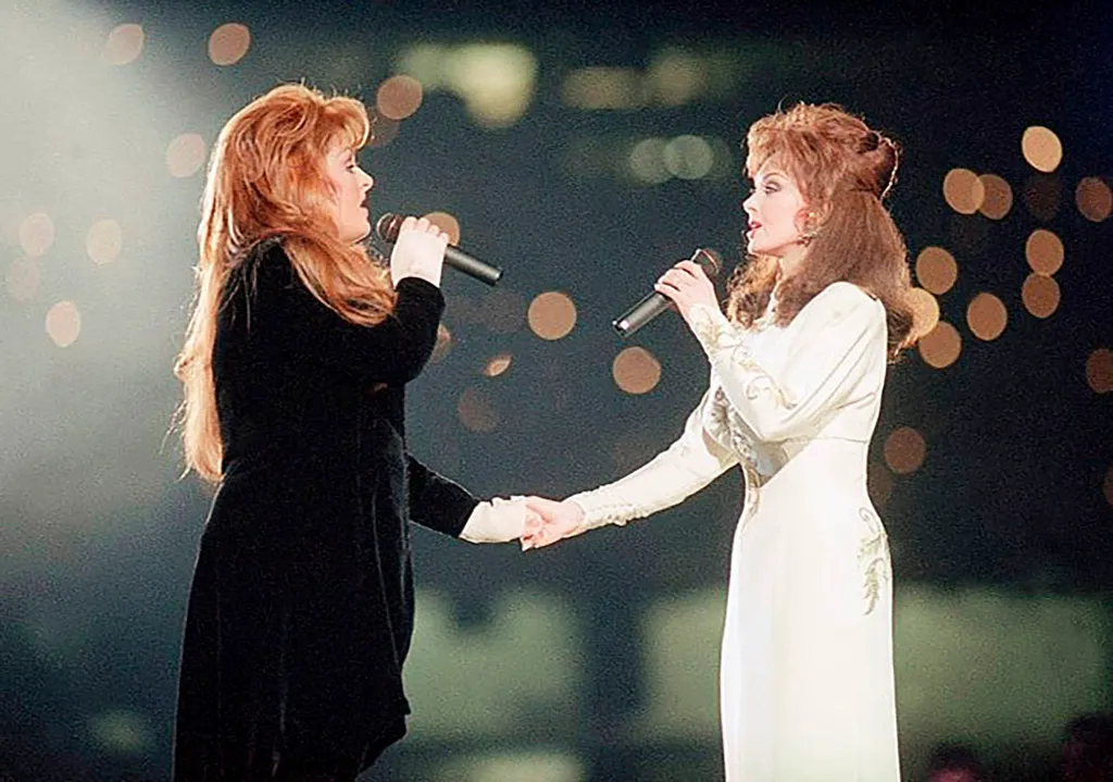 Wynonna and Naomi Judd of The Judds singing into microphones and holding hands on stage.