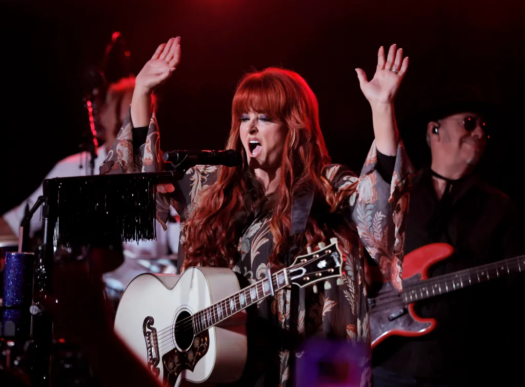 Wynonna Judd performs on stage with her hands raised, singing into a microphone with an acoustic guitar around her neck.