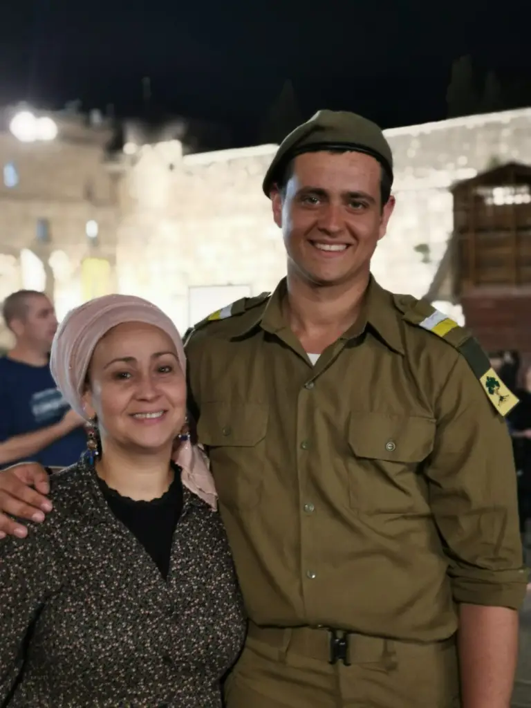 Yasmin Ochayon and her son Eviatar Ochayon Abukasis stand smiling together at night outdoors, with Eviatar in an IDF uniform and beret.
