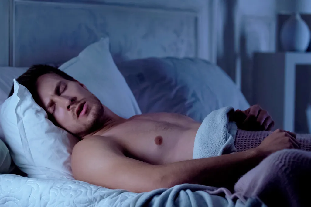 A shirtless young man lying in bed at night, suffering from a nightmare and tossing in his sleep.