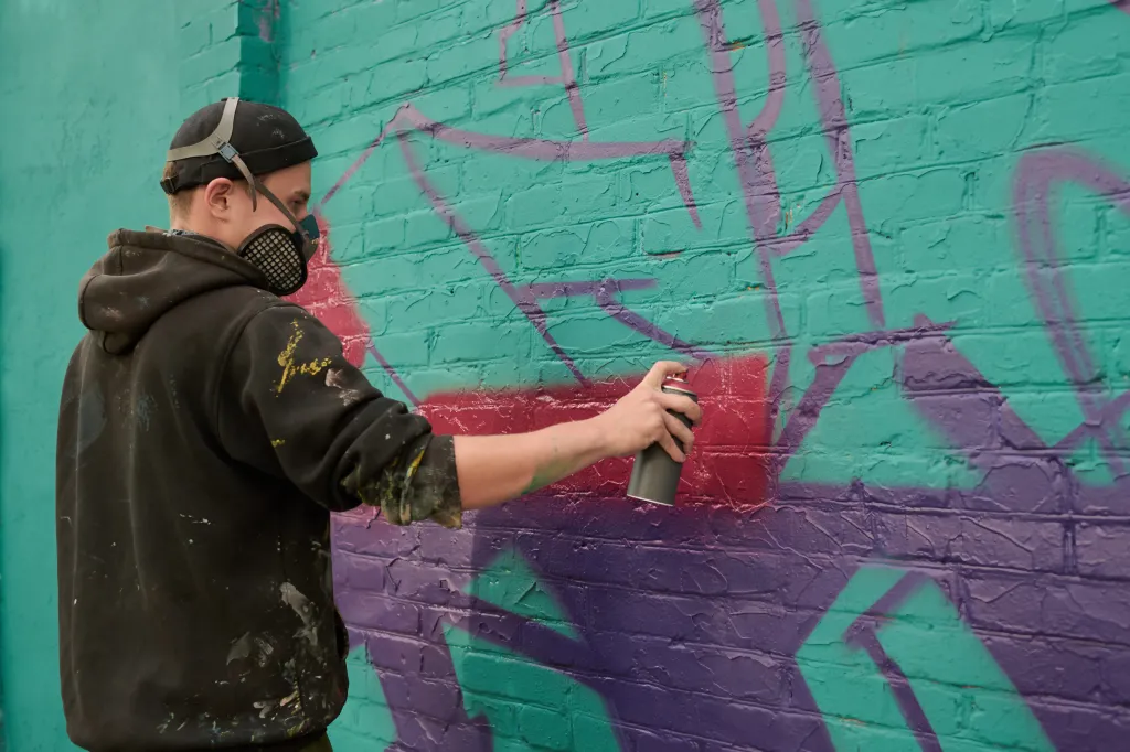 A young man wearing a black hoodie, beanie, and respirator sprays purple paint onto a green brick wall.