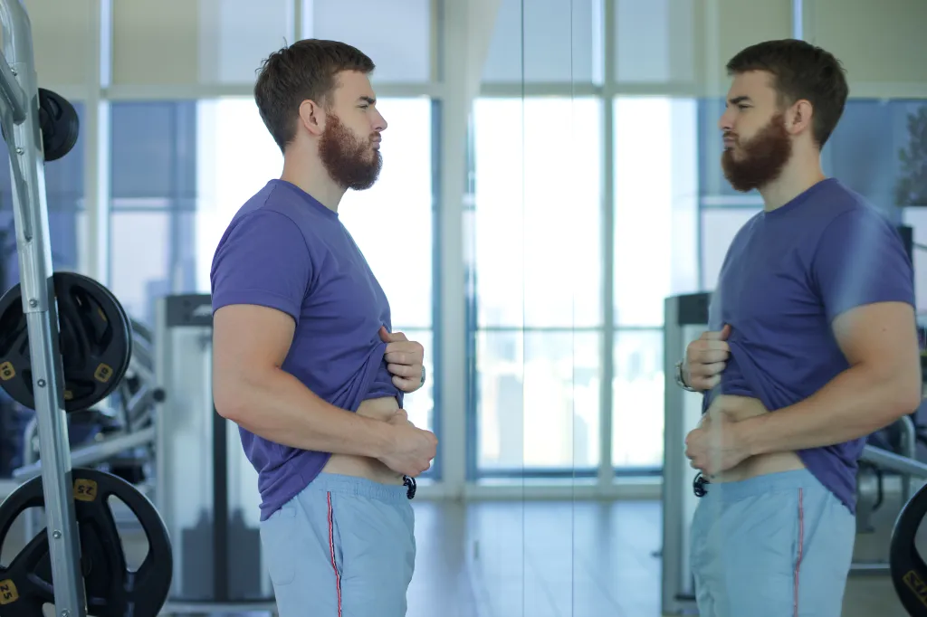 An upset young man in a gym looks at his stomach in a mirror, frustrated with his weight.