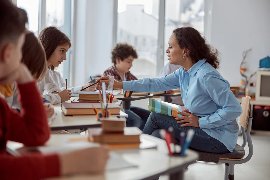 A teacher helps a young student read at her desk while other elementary students study in the background of a bright classroom.