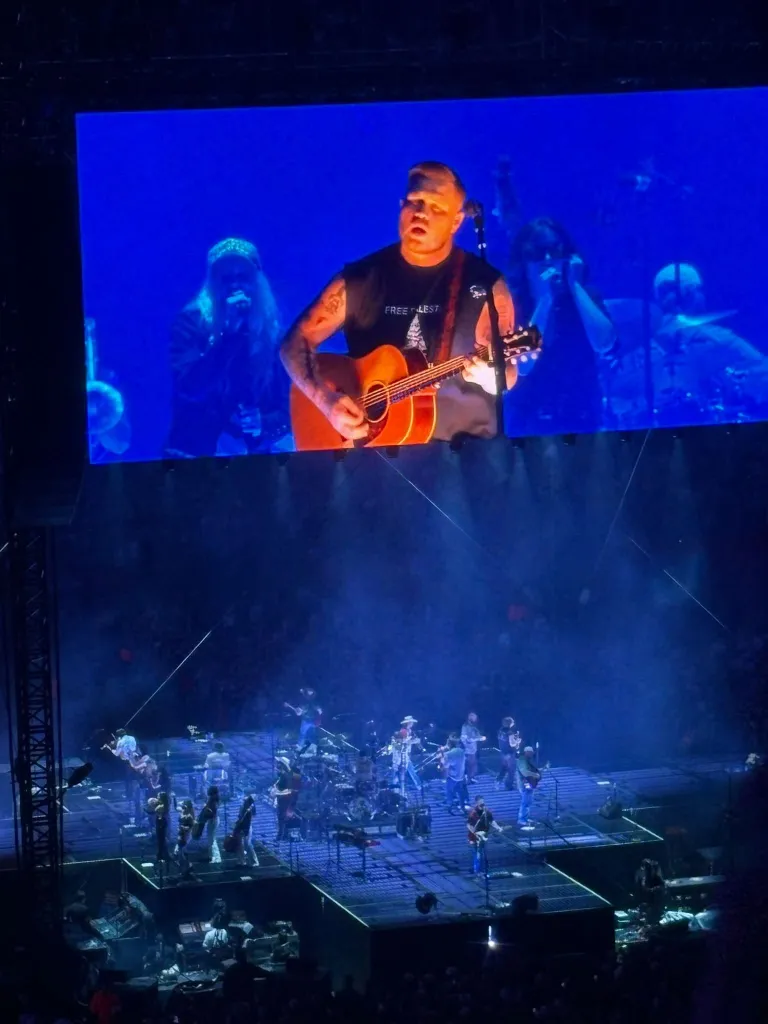 Zach Bryan plays an acoustic guitar on stage, wearing a shirt that reads
