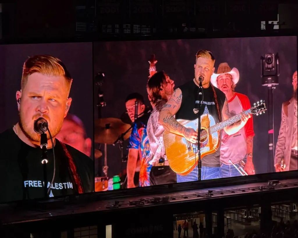 Zach Bryan performs on stage wearing a black shirt that says