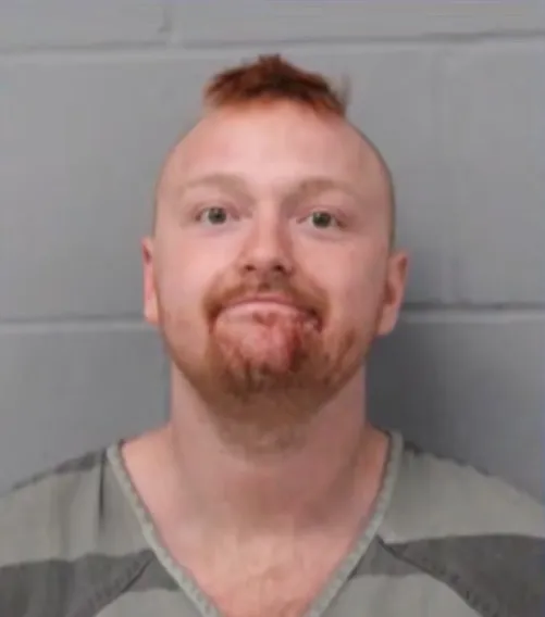 Mugshot of a smiling man with a mohawk and a reddish beard, wearing a striped jail uniform in front of a gray brick wall.