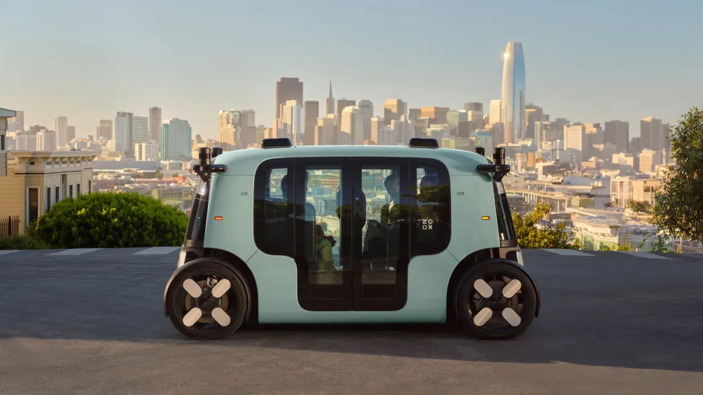 A light-blue Zoox autonomous taxi with a person visible inside, on a street overlooking a cityscape with tall buildings.